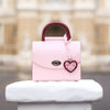 Pink handbag with a heart-shaped charm on a stone pedestal in an urban setting