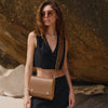 Woman wearing a black sleeveless top and sunglasses, holding a leather cappuccino bag against a rocky background.