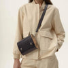Person wearing a beige jacket with a black crossbody bag featuring a wooden tag.